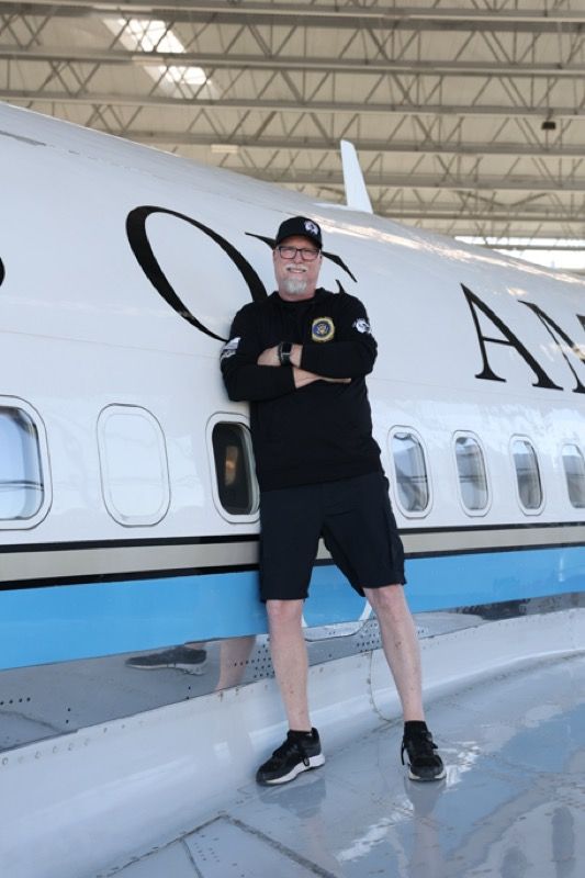 Man in black outfit leans against a white airplane, arms crossed, in a hanger setting.