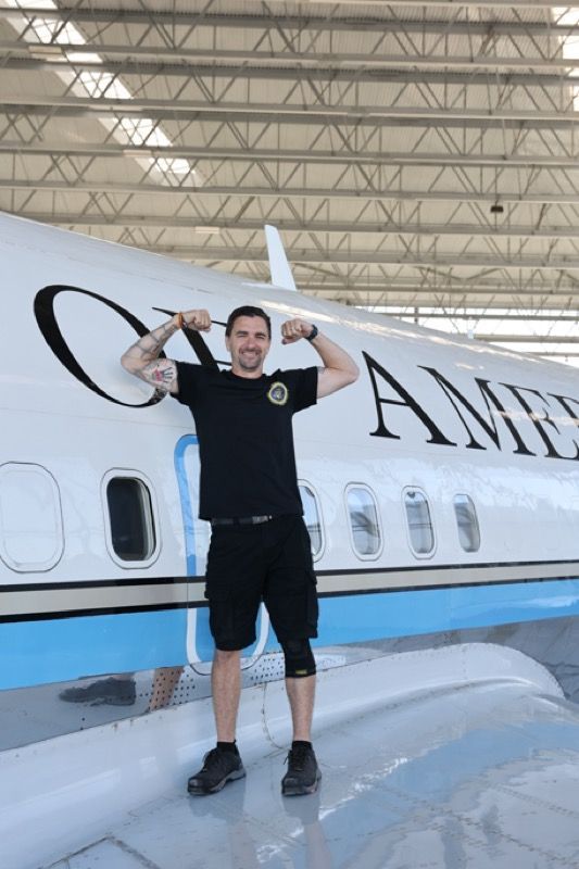 Man flexes arms, stands next to a white airplane, 