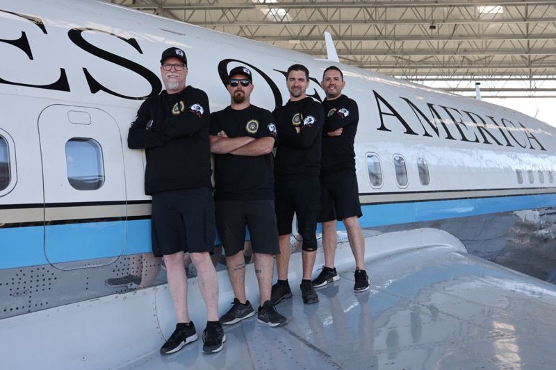 Four men in black pose on a plane wing. The words 