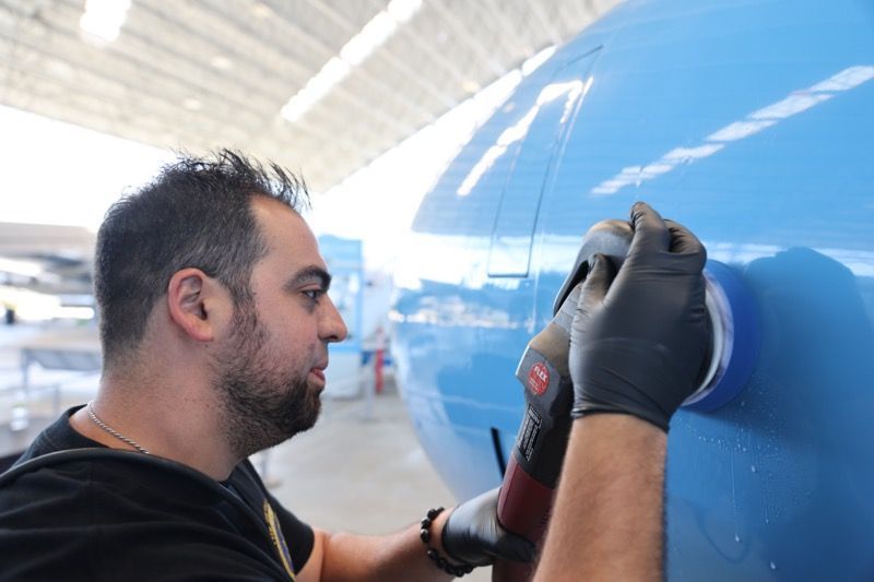Man in black gloves polishing blue airplane fuselage with a power tool in a hangar.
