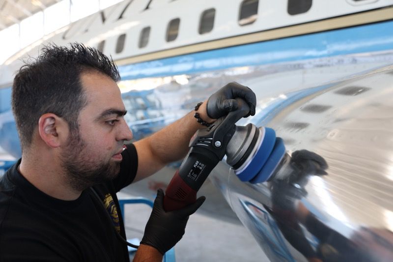 Man polishing an airplane's shiny metal exterior with a power buffer. Black gloves and shirt, blue and white plane.