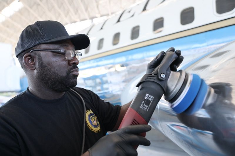 Man polishing an airplane, wearing black gloves, cap, and glasses. The plane has blue accents.