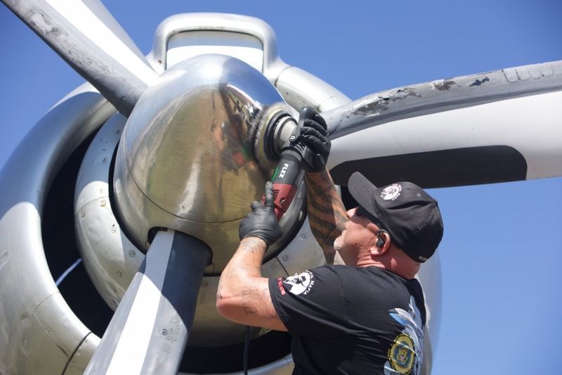 Mechanic working on the propeller of a silver aircraft, outdoors on a sunny day.