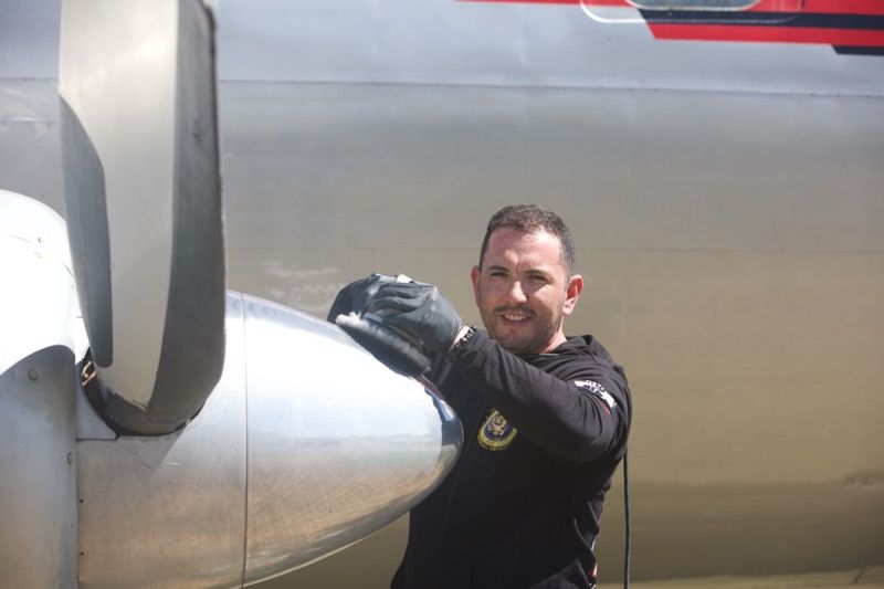 A man in black clothing works on an airplane propeller, smiling outdoors on a sunny day.