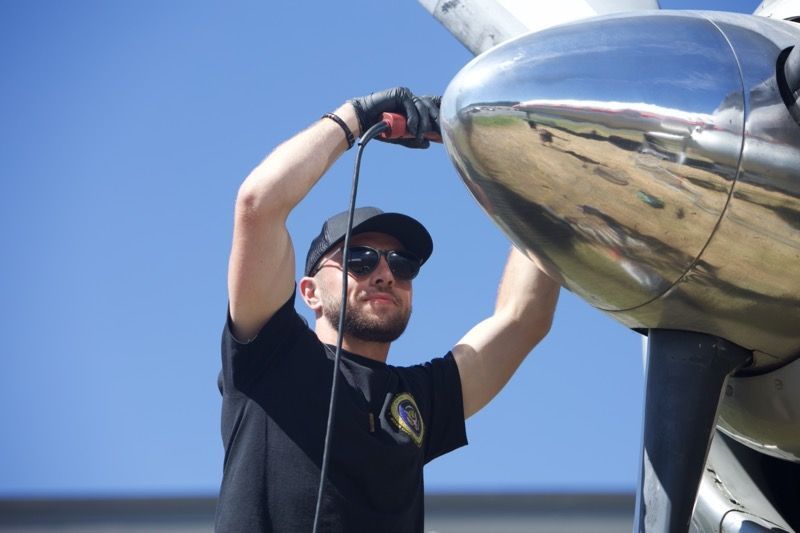 Man in black shirt and cap working on airplane propeller, blue sky.