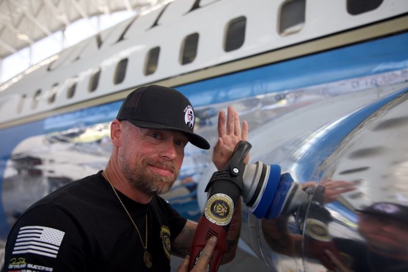 Man in front of an airplane, holding a prosthetic arm. He wears a hat and smiles.