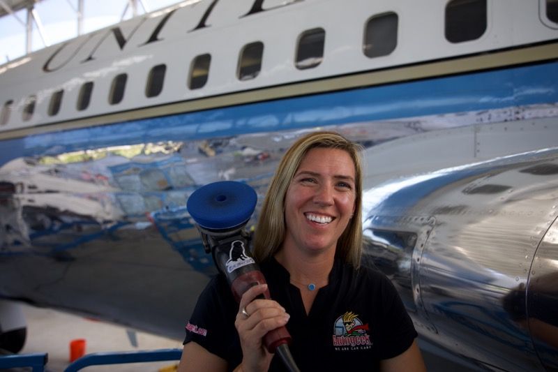 Woman with refueling hose smiles in front of a shiny, blue and silver airplane that says 