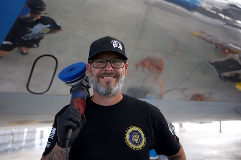 Man with tool smiles in front of an airplane wing; he wears glasses, a black cap, and a T-shirt.