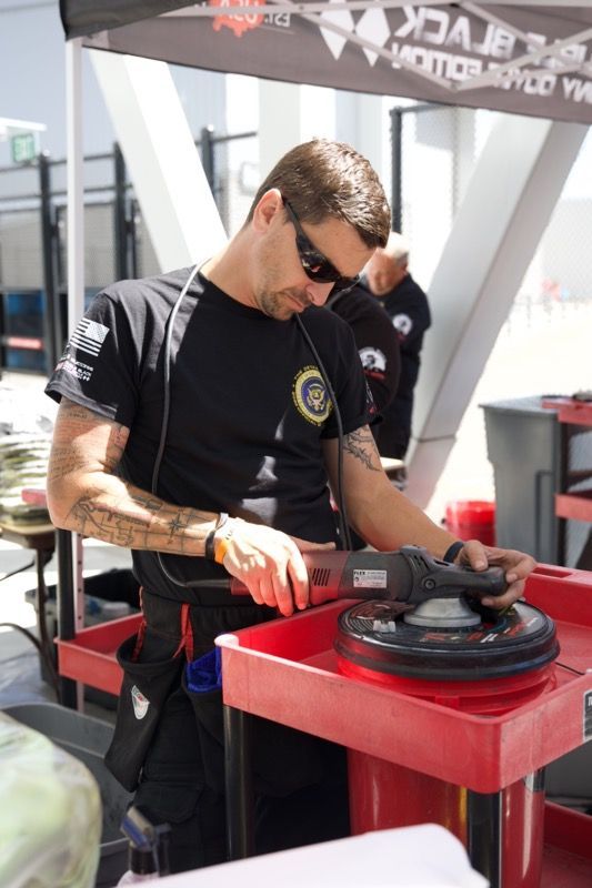 Man polishing a car part, wearing sunglasses and black shirt, outdoors.