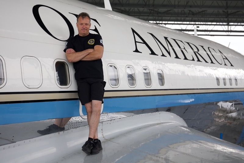 Man in black shorts and shirt leans against a white, blue, and black airplane labeled 