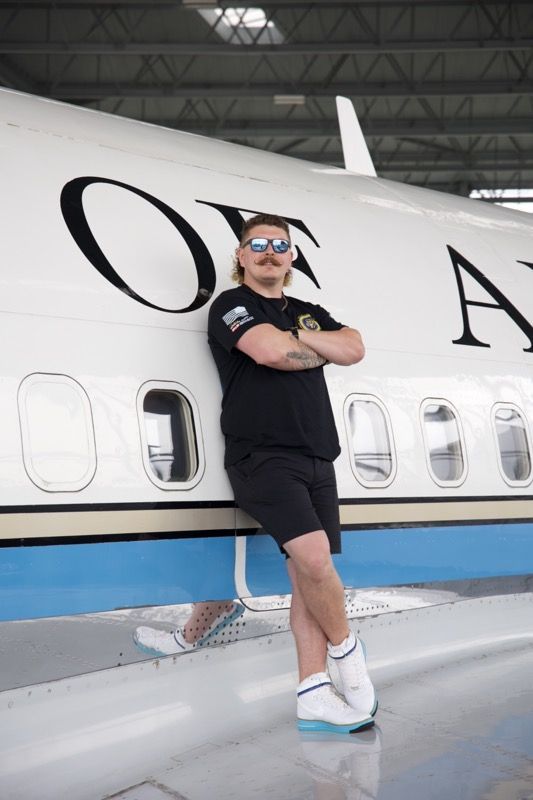 Man with mustache leans against a white airplane, arms crossed, wearing black shirt, shorts, and sneakers.