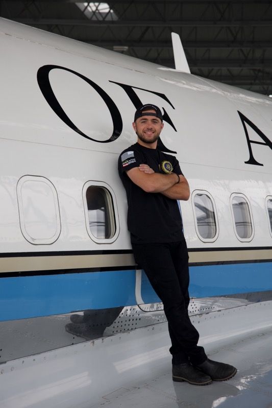 Man in black leaning against the side of a plane, arms crossed. Plane has white fuselage, blue stripe, and 