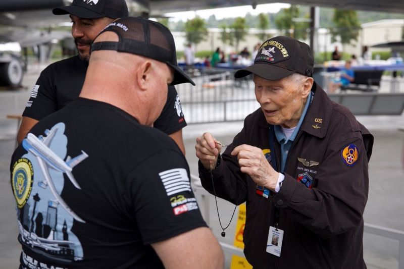 Veteran showing necklace to two men at an outdoor event. He wears a cap and jacket. The men wear black shirts.