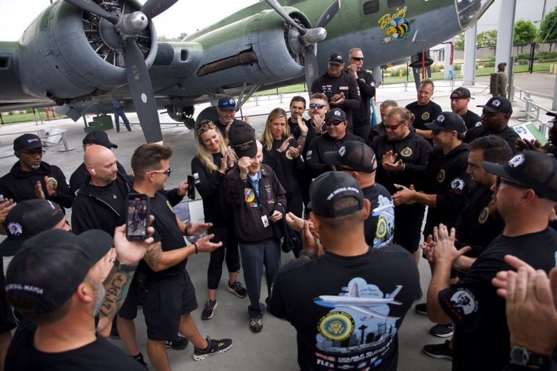 People gathered around a WWII bomber, applauding a woman holding a portrait, likely a memorial.