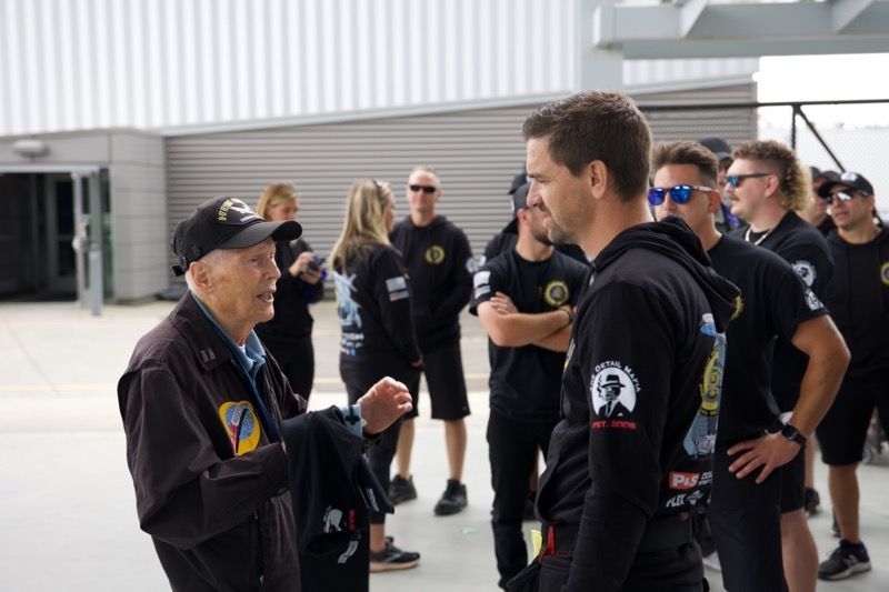 An older man in a cap speaks to a man in black as a group of people look on, outdoors.