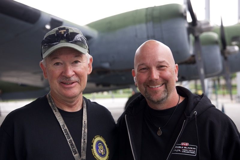 Two men smiling in front of a large, green airplane. One man wears a cap; the other has a shaved head.