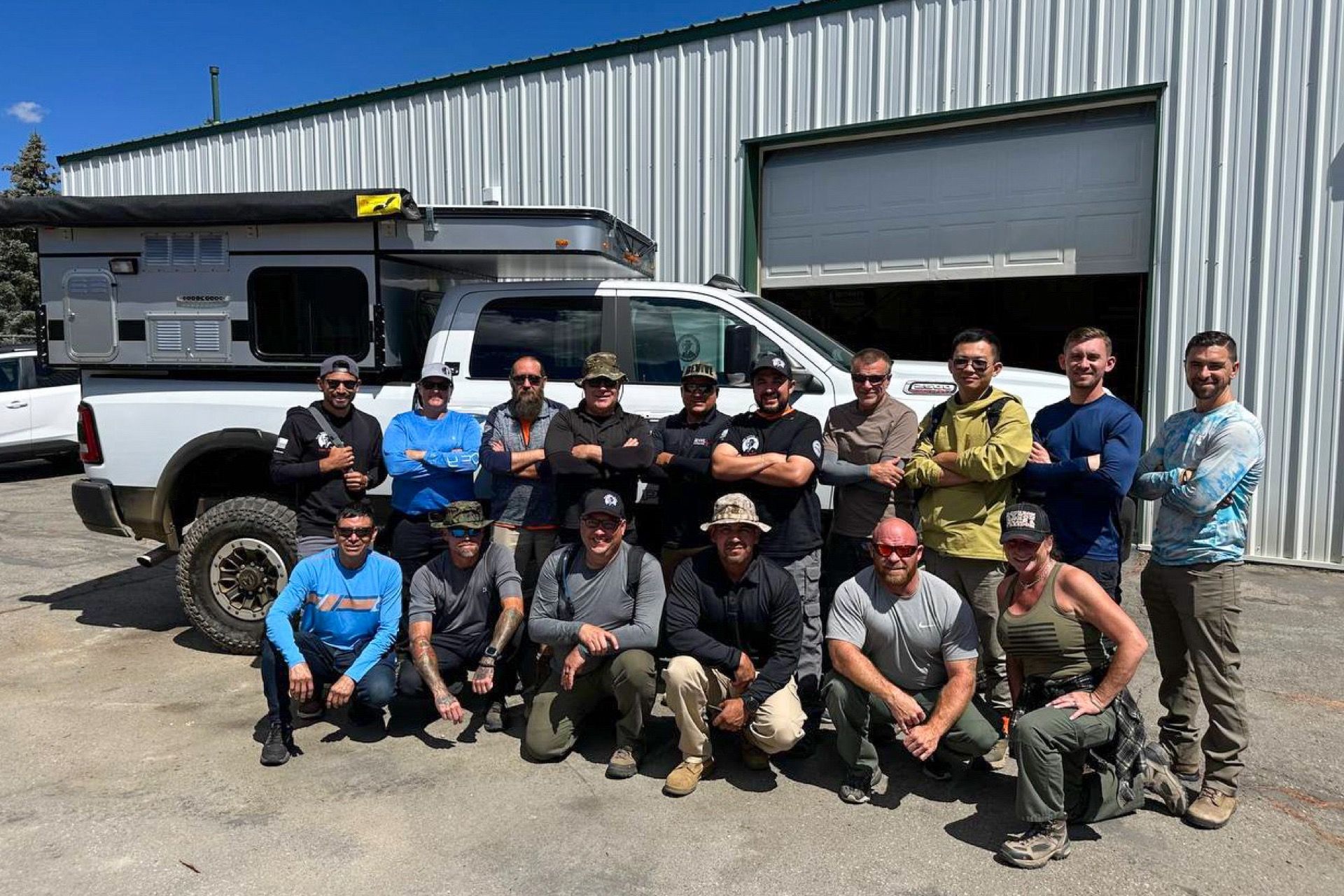 Group of people in front of white truck with camper. Some people are kneeling. Outdoors, sunny day.