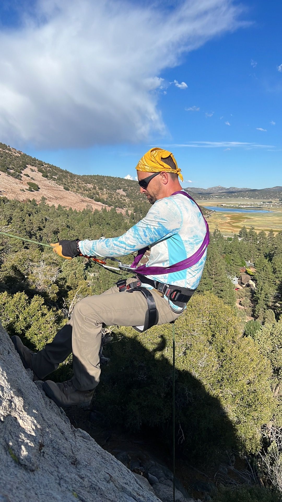 Man rappelling down a rock face outdoors on a sunny day, wearing a harness and gloves.