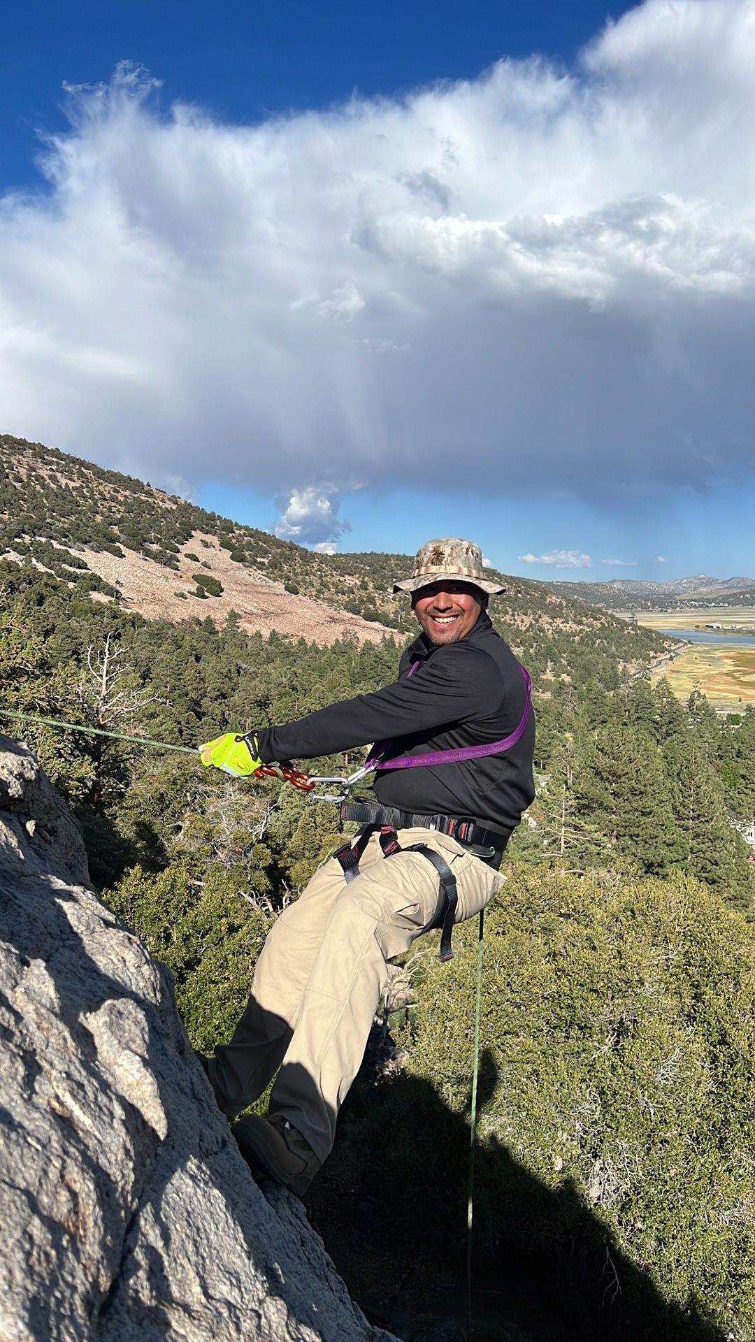 Man rappelling down a rocky cliff face, smiling. Wearing a hat, purple harness, against a mountain landscape and blue sky.