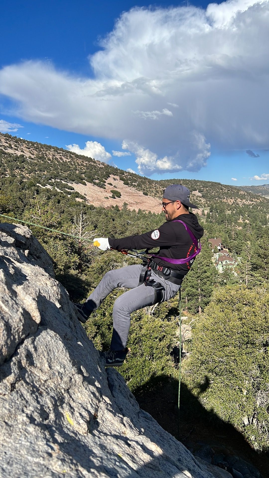 Man rappelling down a rock face, secured with a harness, surrounded by green vegetation under a cloudy sky.