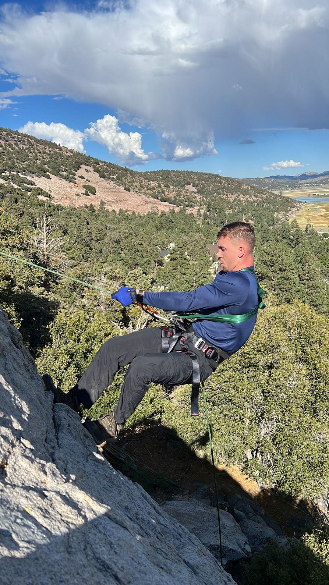 Man rappels down a rock face wearing a harness, blue shirt, and black pants. Outdoors, sunny day.