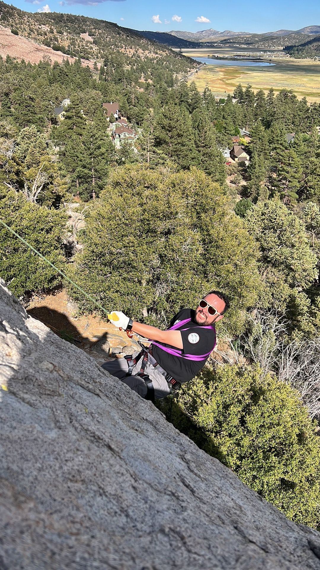 Man climbing a rock face outdoors, wearing sunglasses and a black and purple shirt, sunny day.
