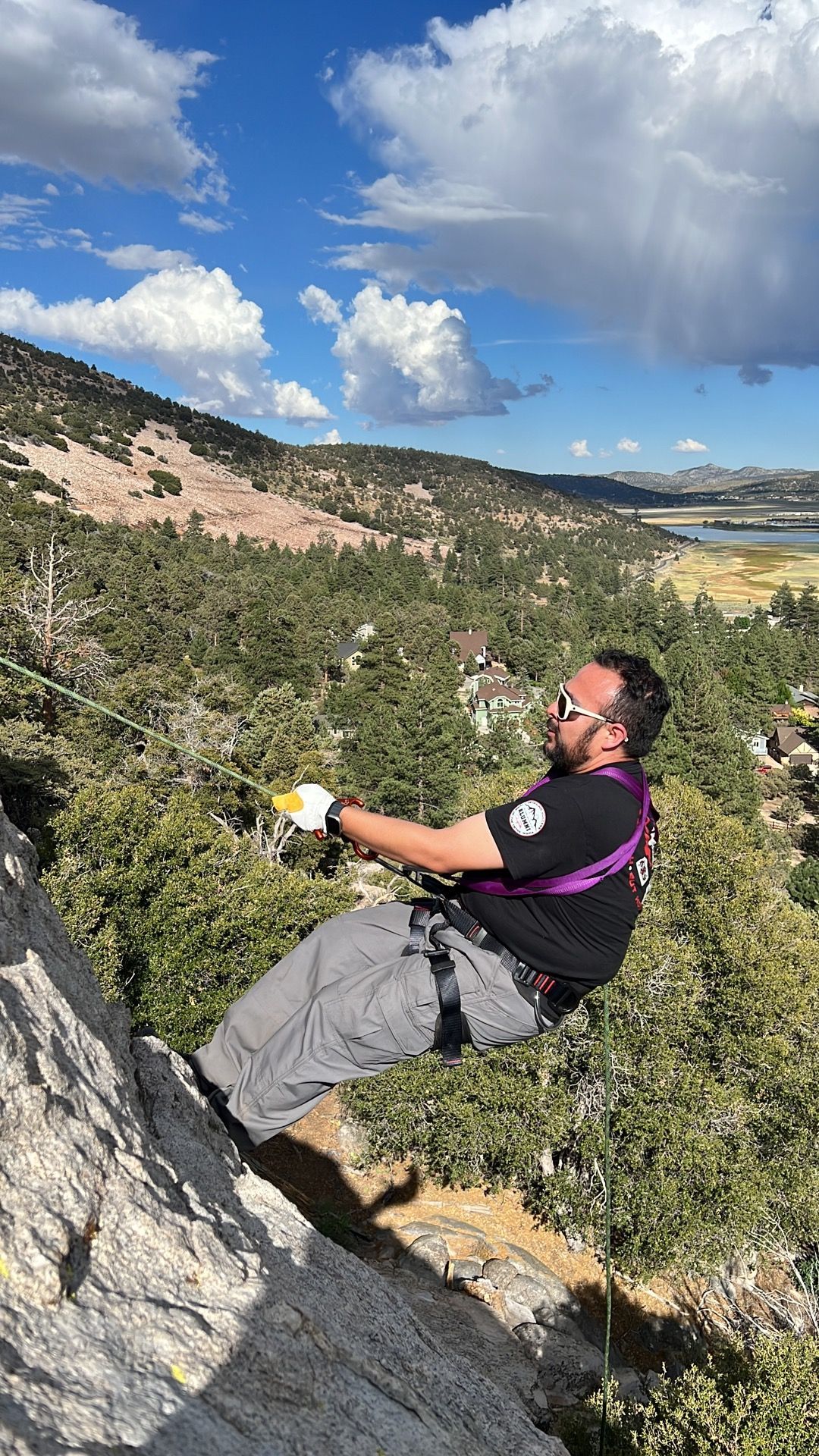 Man rappelling down a rock face, wearing harness and sunglasses, with a mountain and blue sky background.