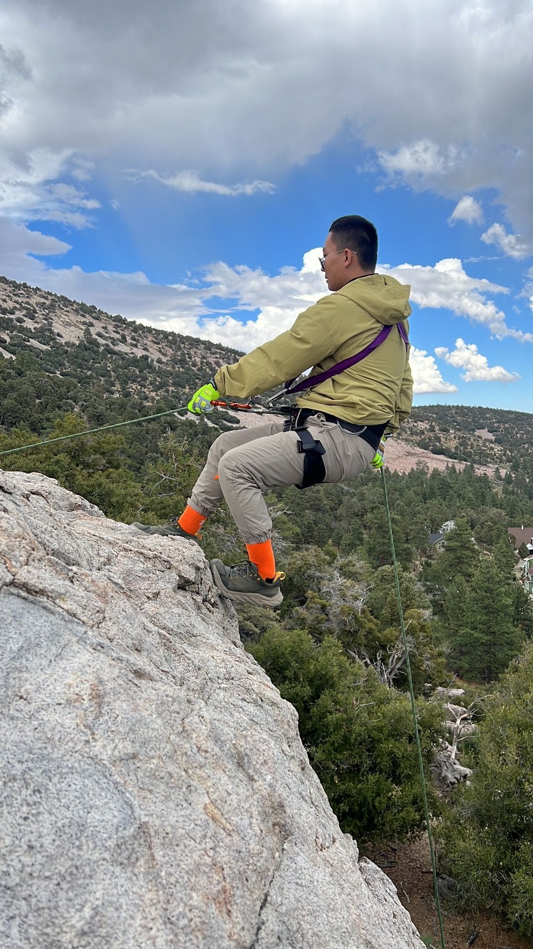 Man rappelling down a rock face, wearing safety gear. Overcast sky, forest backdrop.