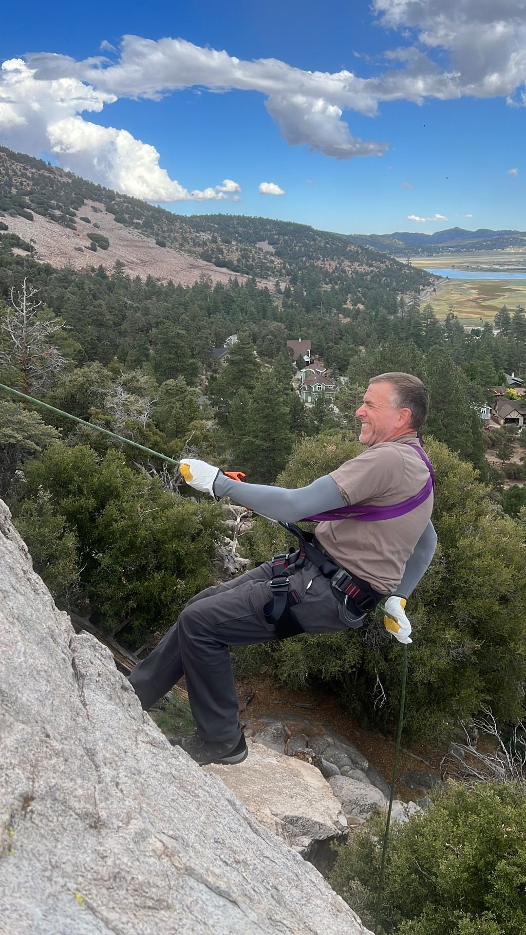 Man rappelling down a rocky mountainside, smiling, with a harness and rope. Sunny, scenic view.