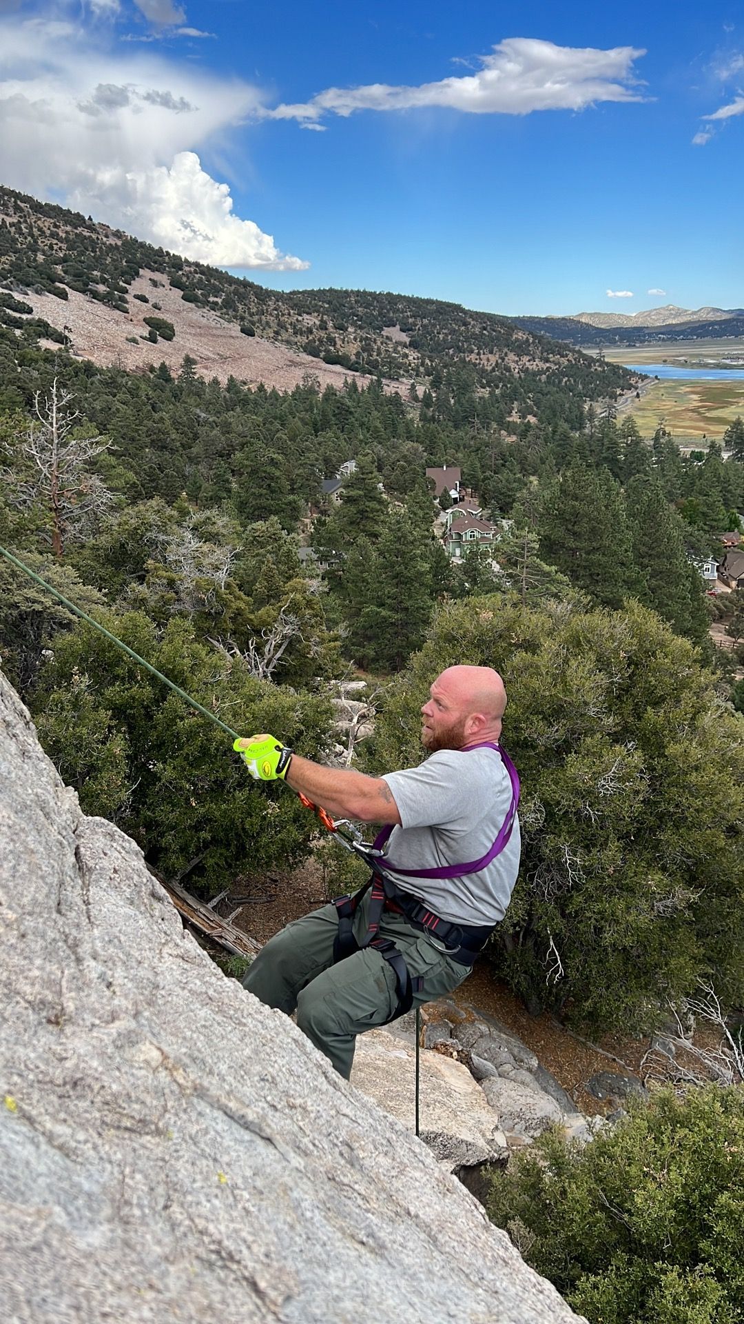 Bald man rappelling down a rock face, wearing a harness, with trees and a lake in the background under a blue sky.