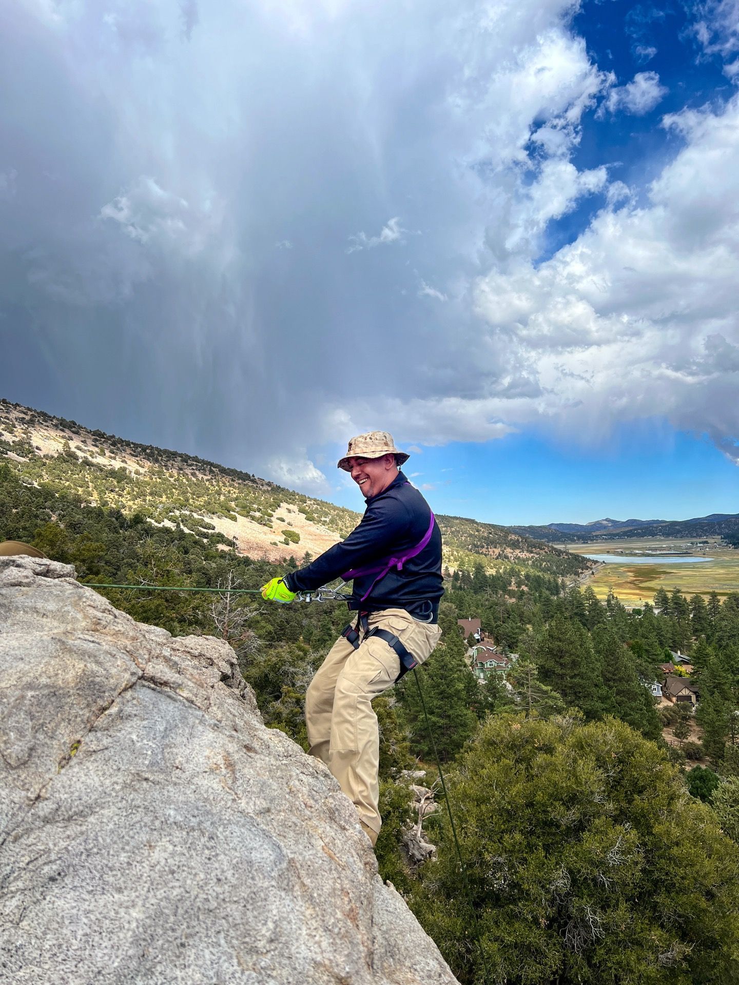 Man rappelling down a rock face, smiling. He's wearing a harness, hat, and jacket. Mountainous landscape, cloudy sky.