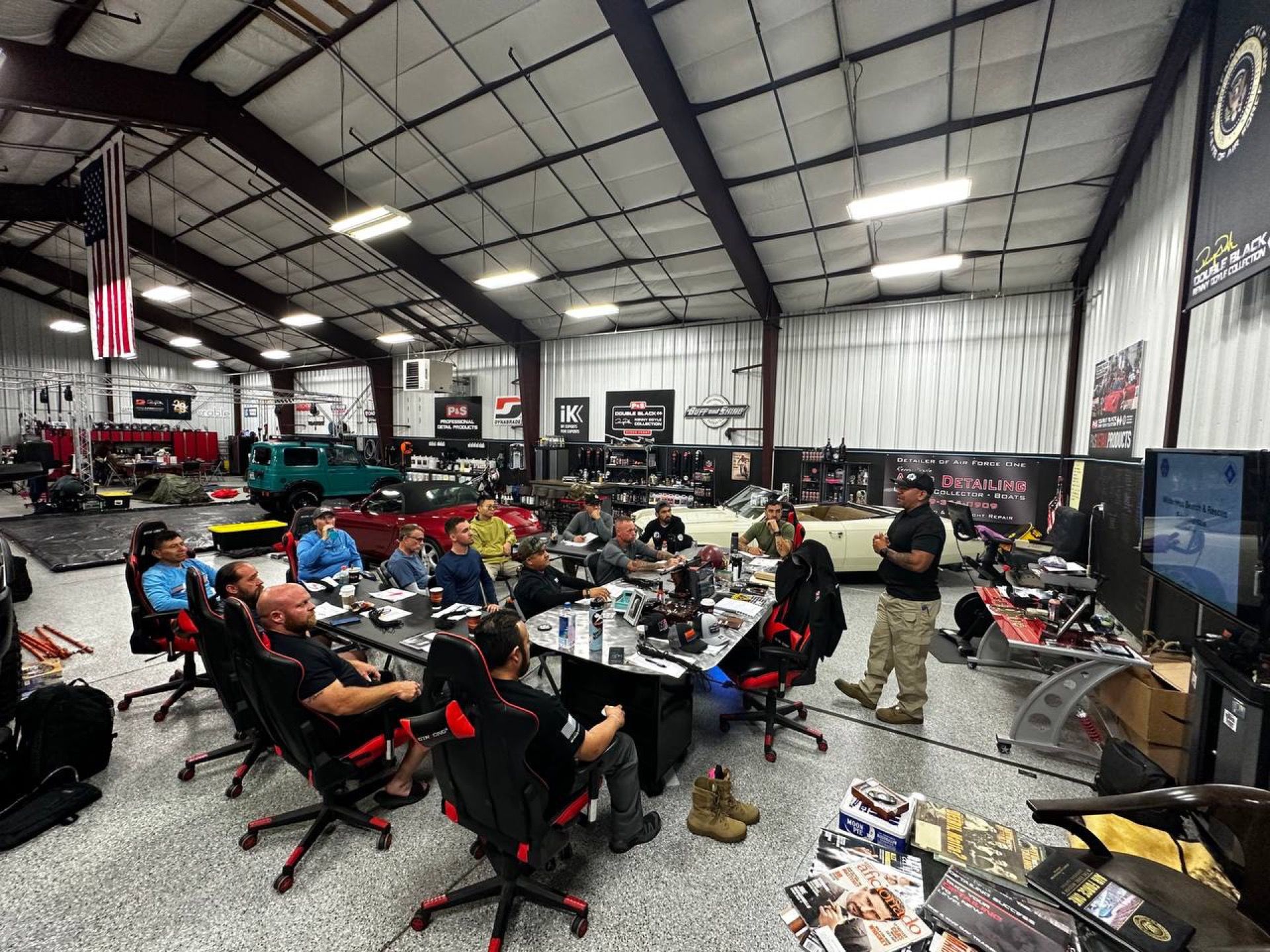 Group of people in a large workshop, gathered around a table for a meeting. American flag hangs in the background.