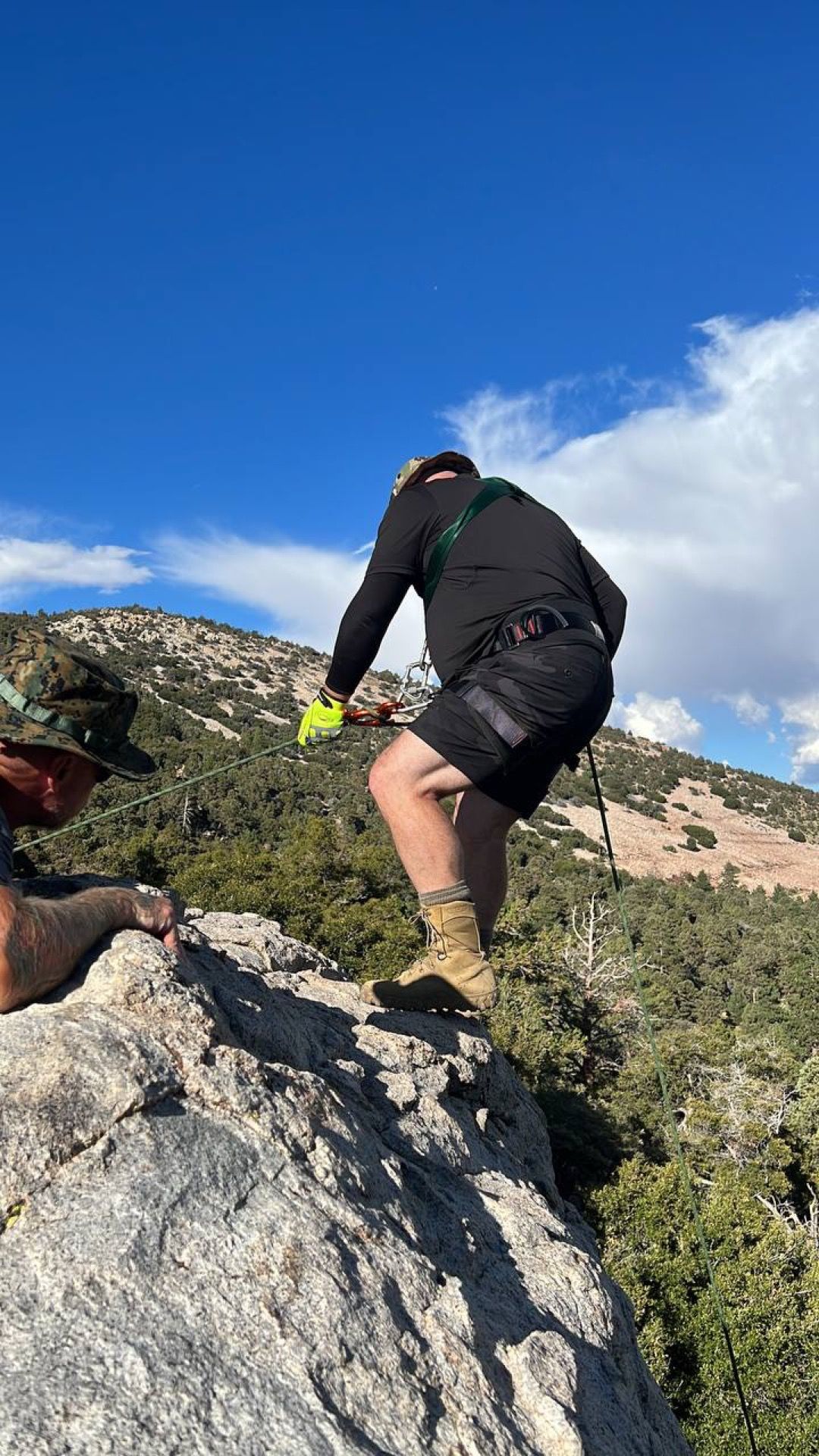 Man in black clothing, gloves, and boots climbing rocky terrain on a sunny day with blue sky.