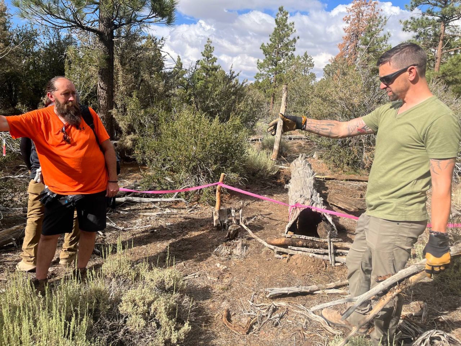 Two men surveying a trail in a forest, marked with pink tape. One points; the other gestures.