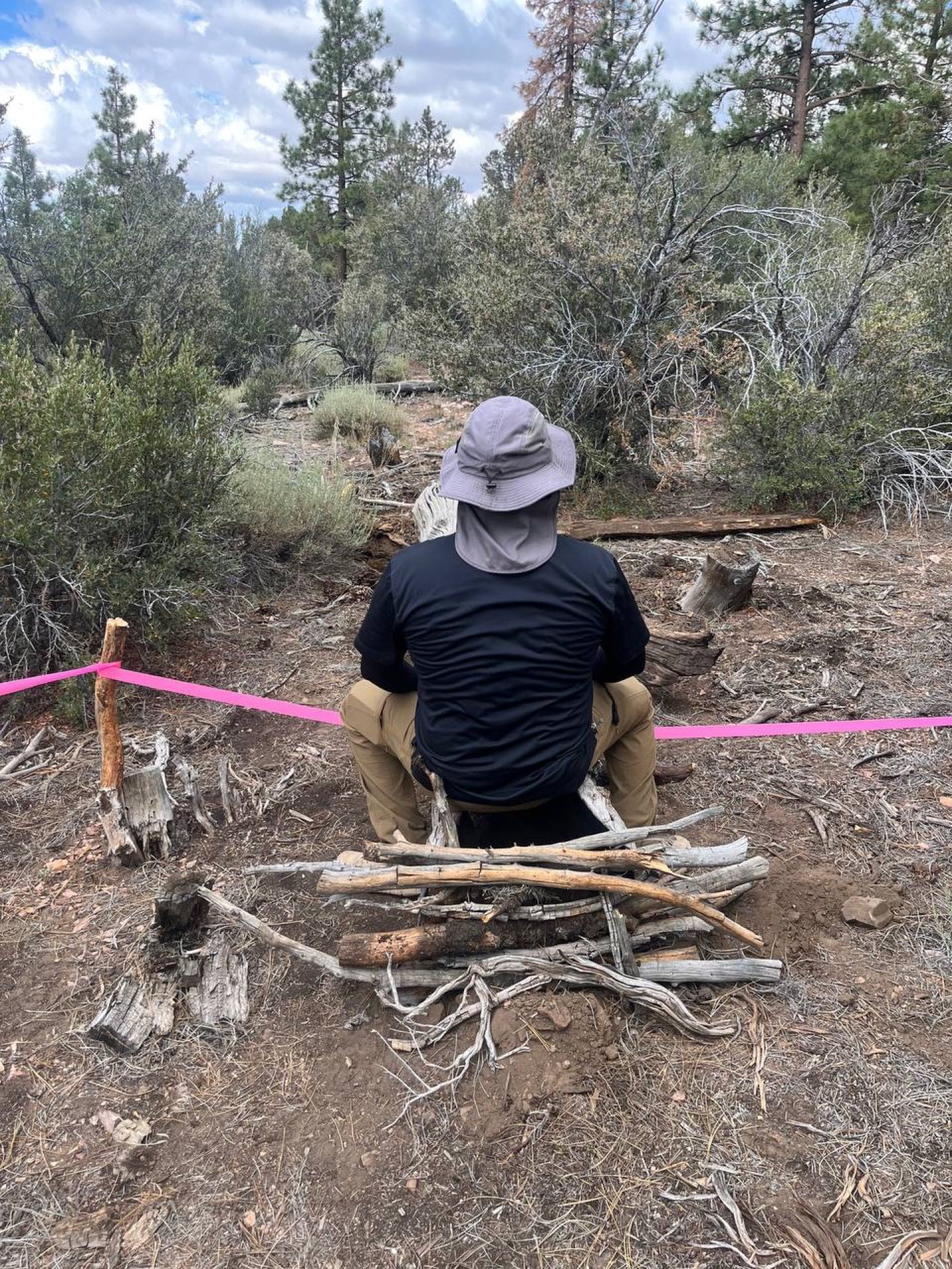 Person in hat squatting near a makeshift campfire, pink tape border, set in a wooded area.