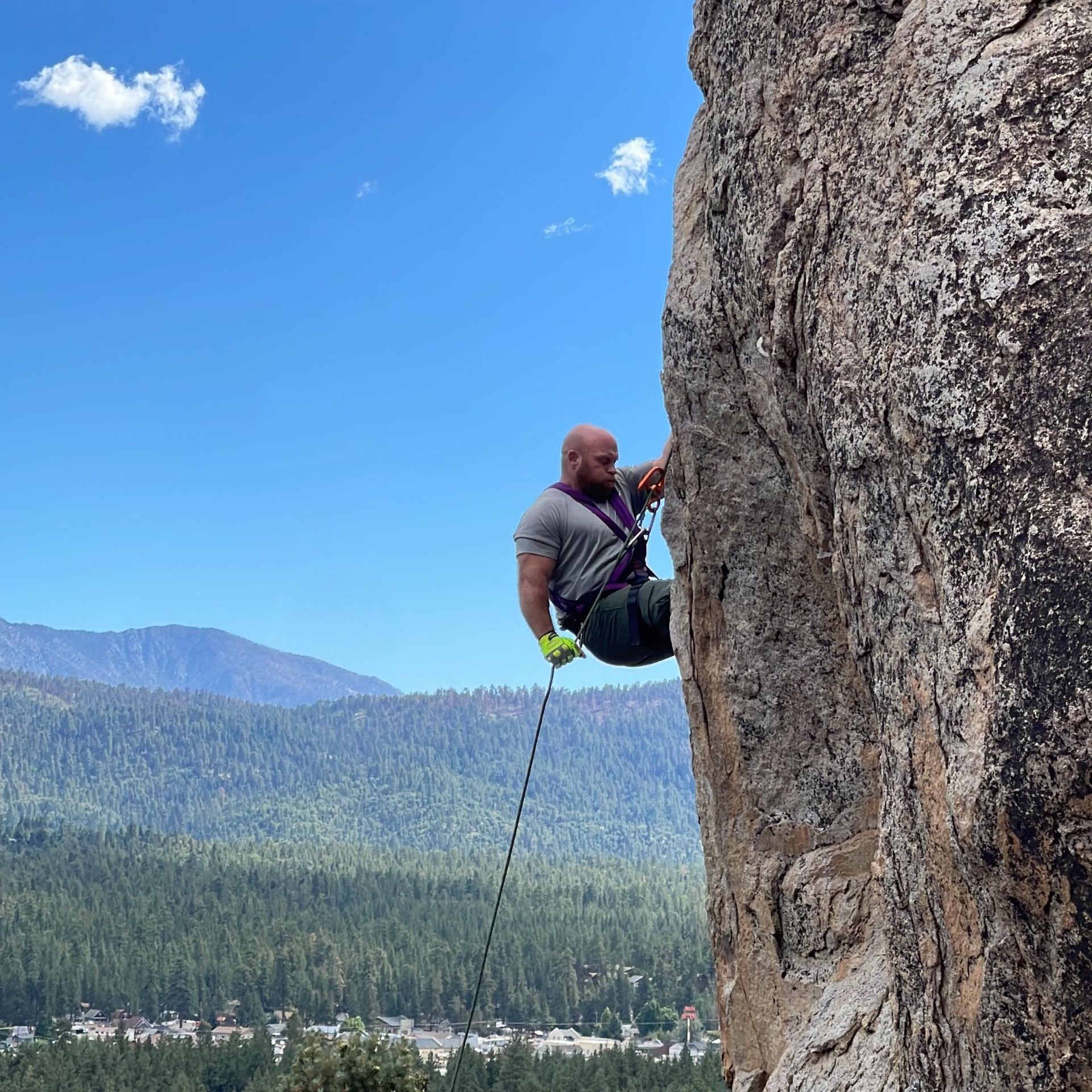 Man rappels down a rock face, wearing safety gear. Blue sky, green trees, and mountains in the background.