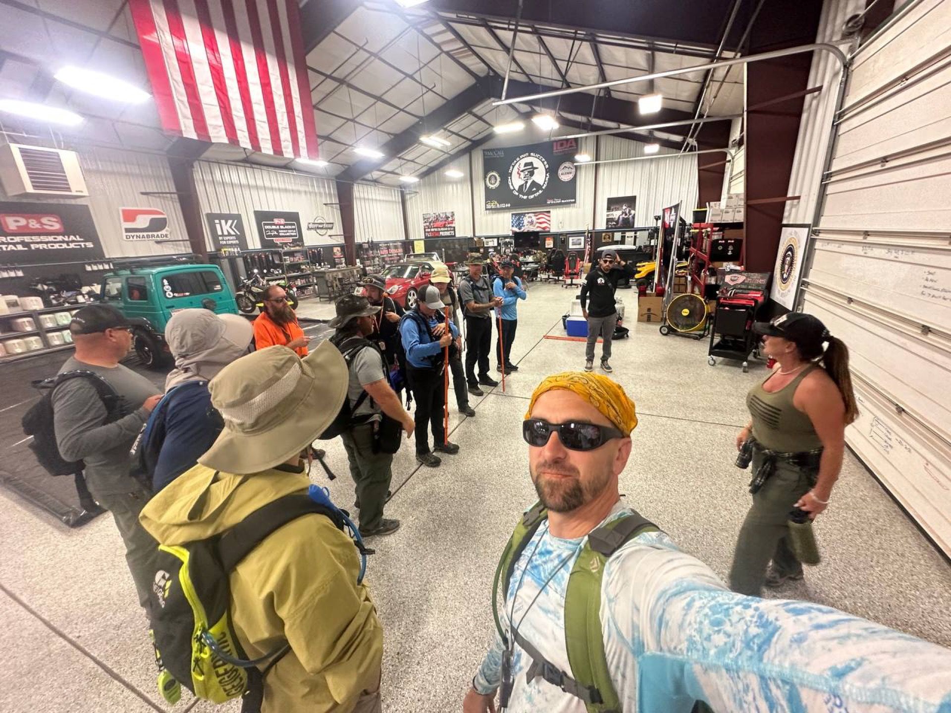 Group of people in a garage-like space, some wearing backpacks, listening to a man, American flag in background.