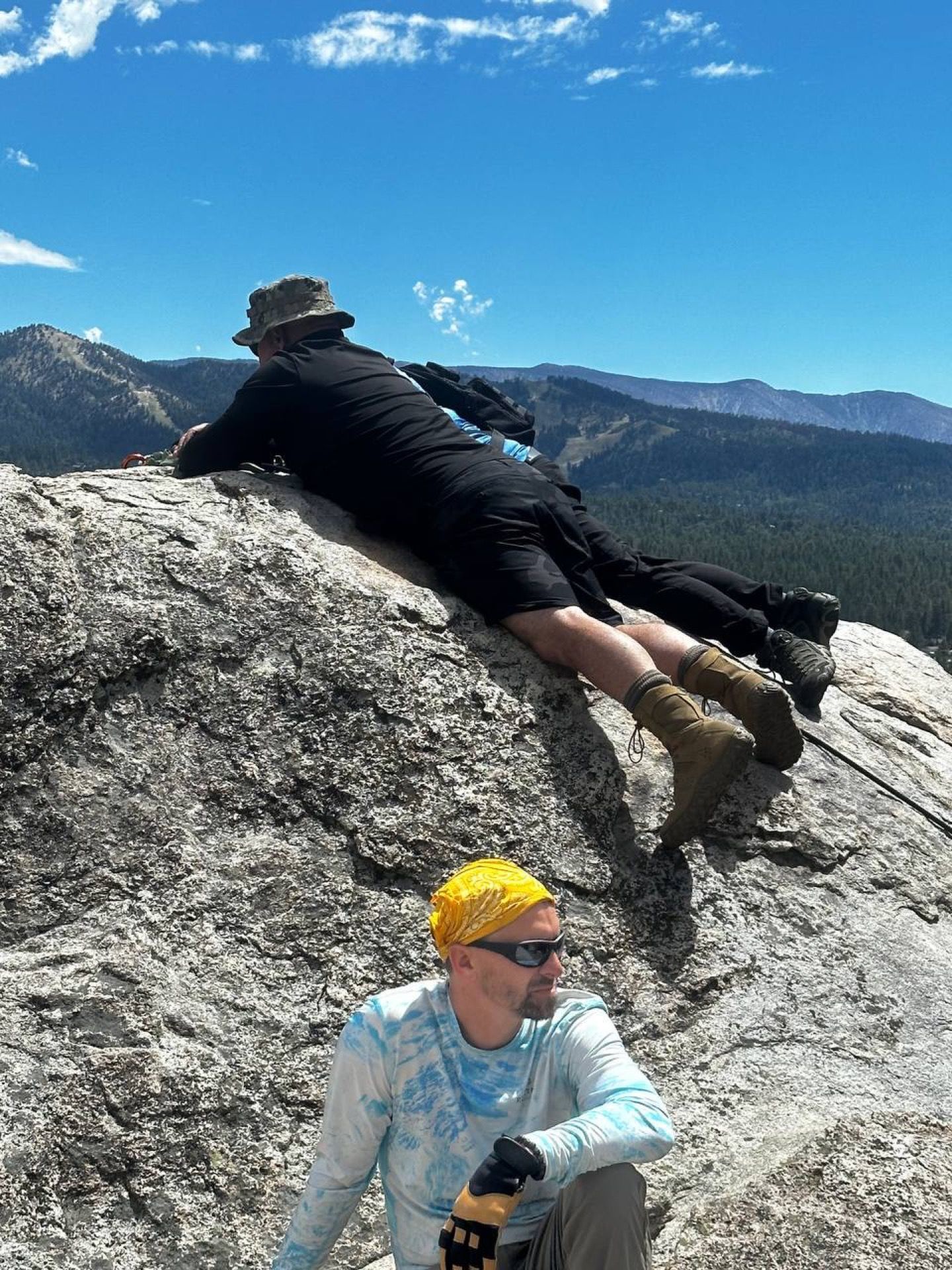 Two people on a rocky mountain summit; one in a hat gazes out, the other rests; blue sky and mountains.