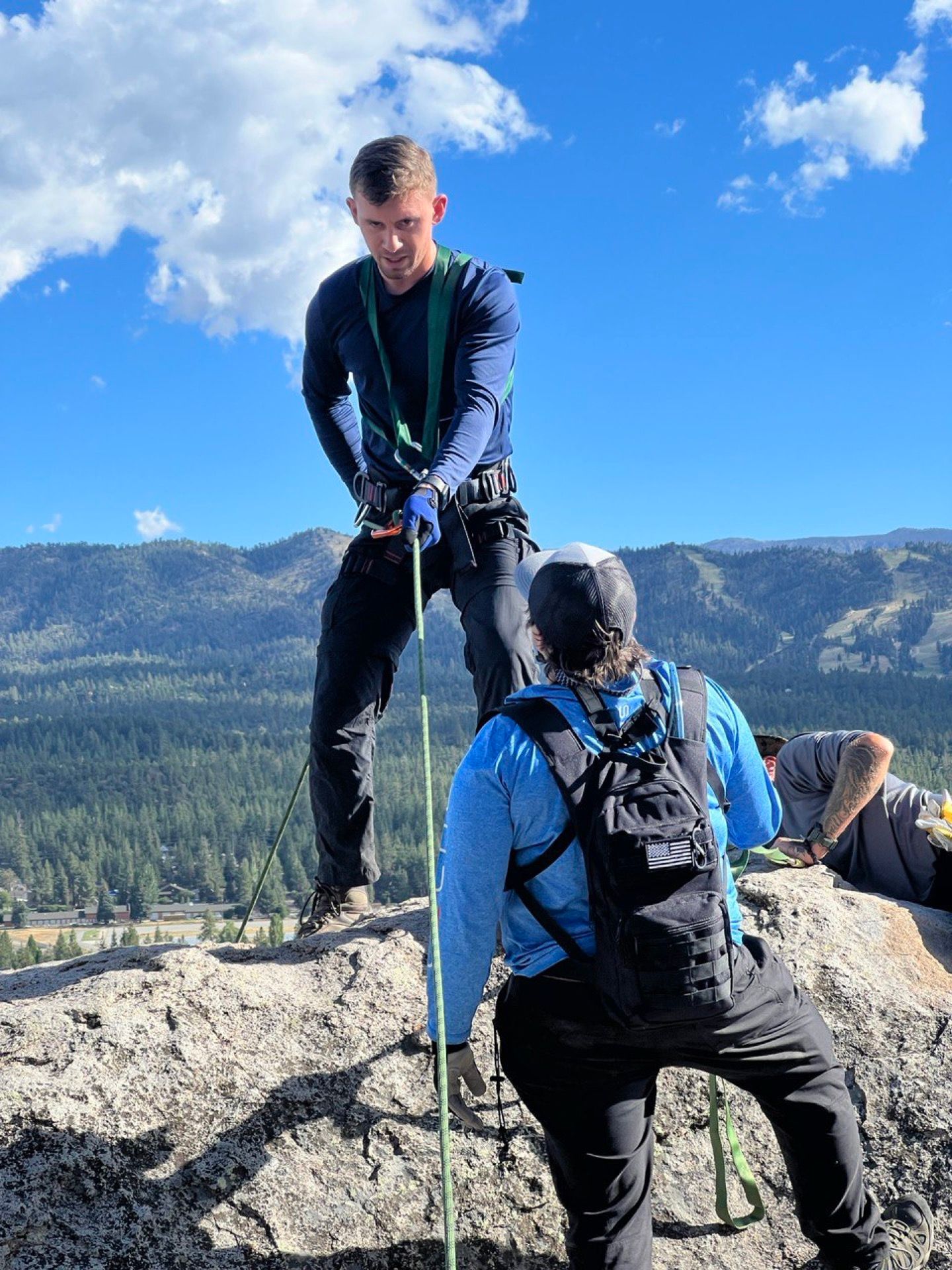 Two people on a rocky cliff face, one rappelling, the other holding the rope. Mountain landscape in the background, blue sky.