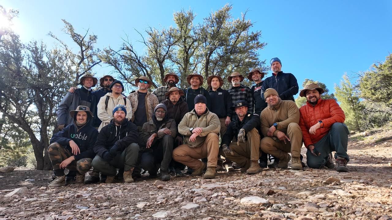 Group of people pose in a mountain setting, most wearing hats. Sunny, blue sky.
