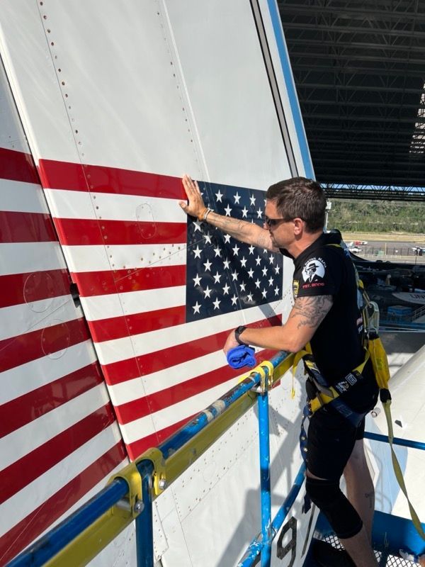 Man in harness adjusts a large American flag on the side of a structure. Outdoors, sunny.
