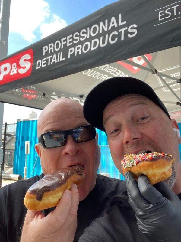 Two men eating donuts, one with chocolate frosting, the other with sprinkles, outdoors under a 