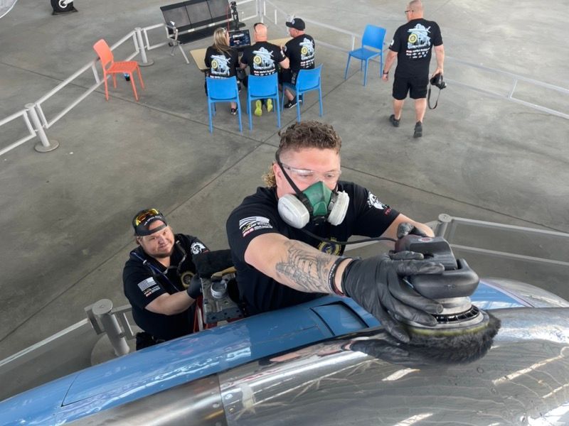 Two men polishing a metal vehicle outdoors. One wears a respirator and goggles. Others watch from chairs.