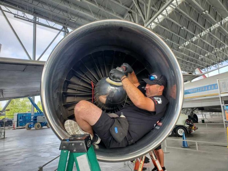 Man polishes the inside of a jet engine while sitting inside. He wears black and uses a power tool.