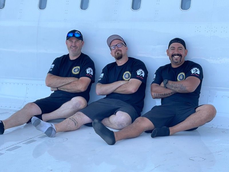 Three men in black shirts, arms crossed, seated on white surface.
