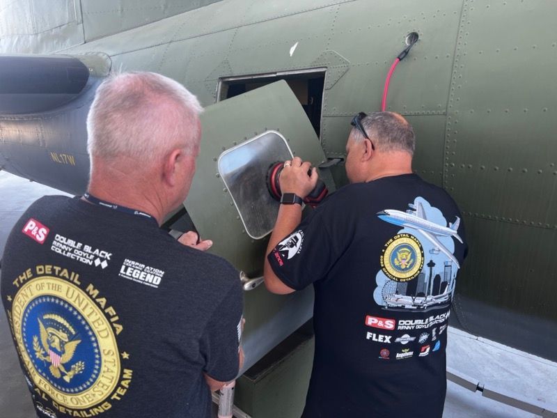 Two men working on the side of a dark green aircraft. One sands a panel, and the other watches.