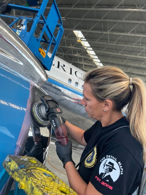 Woman polishes an airplane with a power tool, wearing a black shirt and gloves, inside a hangar.
