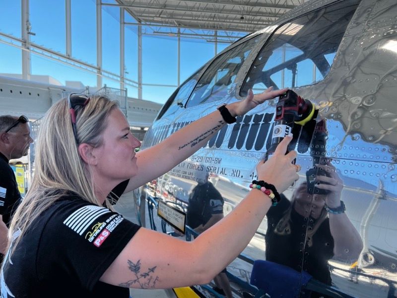 Woman inspecting a silver airplane's cockpit. She wears a black shirt and sunglasses, in an outdoor setting.