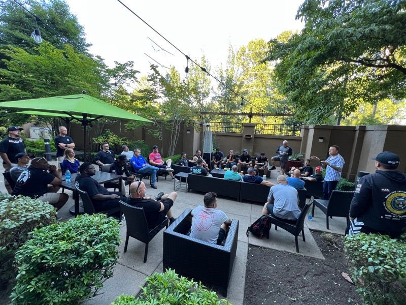 Outdoor gathering of people seated and standing, listening to a man speaking. Trees and green umbrella visible.