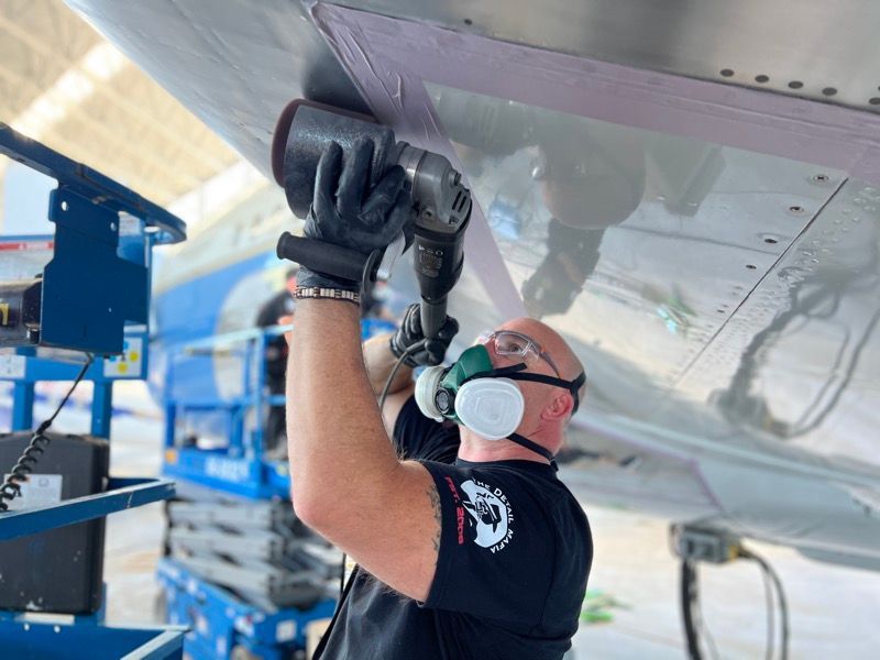Mechanic working on an airplane wing, using a sander, wearing safety gear in a hangar.
